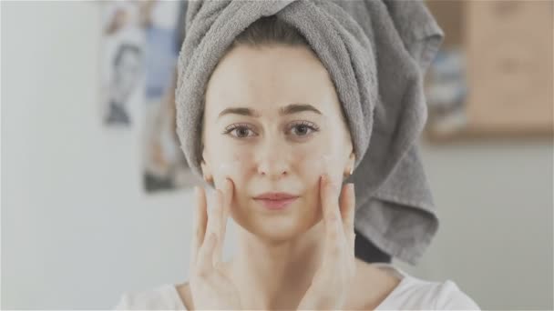 Beautiful young woman with towel on head applying cream on face and look at the camera - Footage, Video