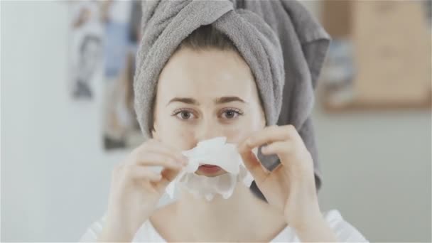 schönes Mädchen mit Handtuch auf dem Kopf zieht feuchtigkeitsspendende Gewebemaske vor dem Spiegel aus - Filmmaterial, Video