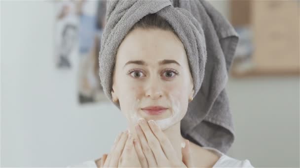 Mujer con toalla en la cabeza aplicando máscara cosmética y mira a la cámara
 - Imágenes, Vídeo
