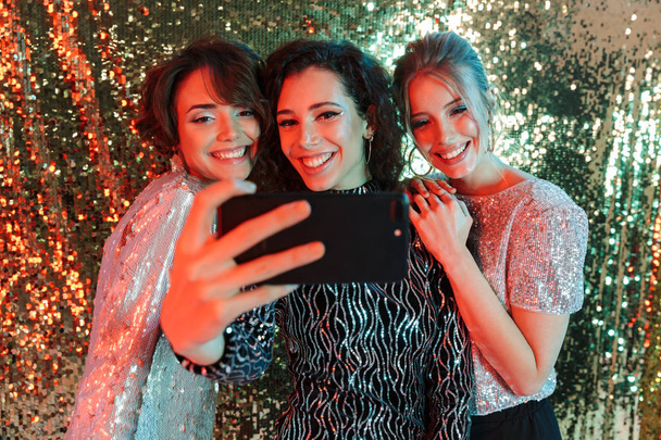 Beautiful young happy women friends posing over glitter wall background take a selfie by phone. - Foto, imagen