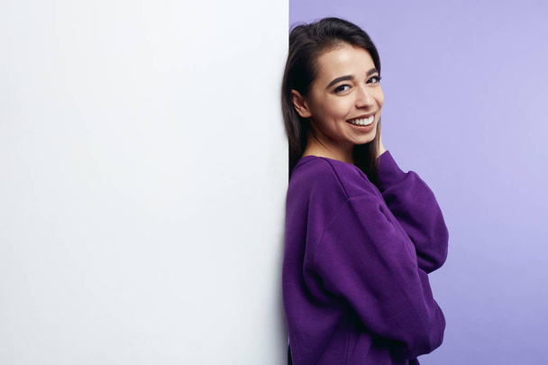 Kopírovat prostoru portrét okouzlující atraktivní dívka, stojící vedle prázdné bílé billboard stěna izolovaná fialové pozadí. - Fotografie, Obrázek