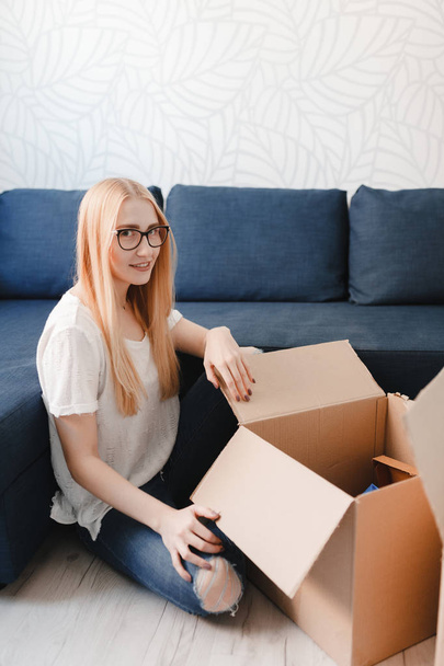 Moving house: Young woman moving into new apartment holding cardboard boxes with belongings, blonde girl sittng near sofa with boxes - Foto, Imagen