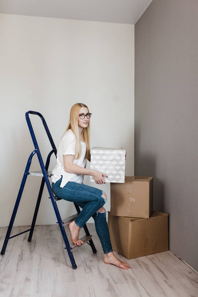 young woman sitting on ladder in empty flat with cardboard boxes during relocation at new home - 写真・画像