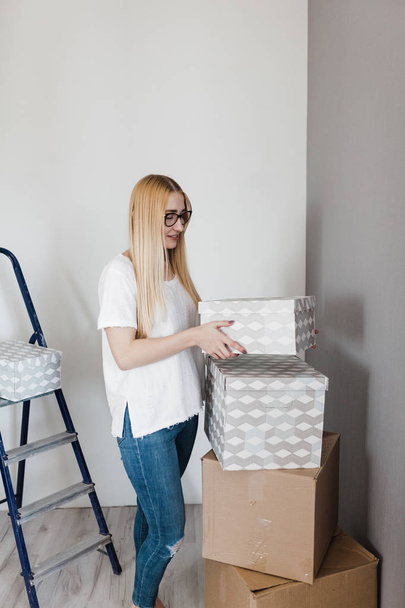 young blonde happy woman in whitet-shirt standing on empty wall with some moving boxes - Foto, afbeelding