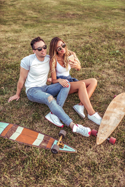 Man woman relationship summer city sitting grass, taking pictures phone, taking selfies. Longboard skateboard, sunglasses. Emotions joy fun, relaxing fresh air weekends. Online video call application. - Photo, image
