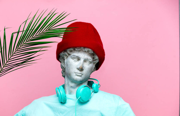 Античний бюст чоловічої статі в капелюсі з навушниками і гілкою долоні на рожевому фоні. Готовий до літніх канікул
 - Фото, зображення