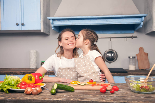 Bonito filha beija mãe na bochecha enquanto cozinha em uma bela cozinha azul
 - Foto, Imagem
