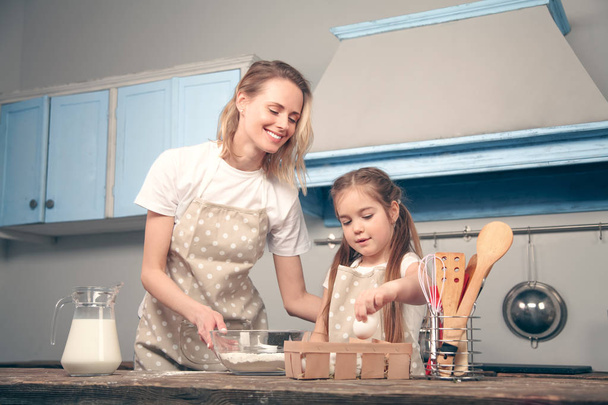 Mutter und Tochter in der Küche kochen Mafins. eine Tochter hält ein Hühnerei in der Hand, das zum Mehl hinzugefügt wird - Foto, Bild