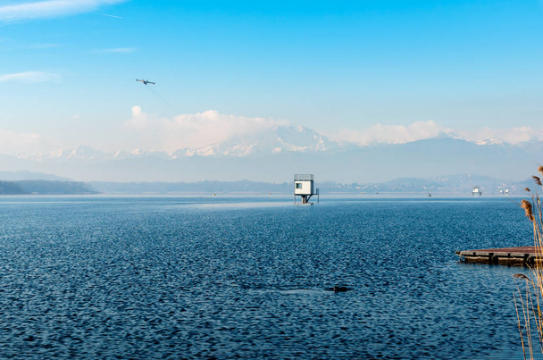 Τοπίο της λίμνης Varese σε μια ημέρα του χειμώνα, με το αεροπλάνο που πετούν πάνω από τα βουνά με το χιόνι που καλύπτει κορυφές στο παρασκήνιο. - Φωτογραφία, εικόνα