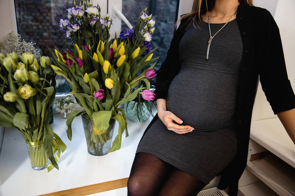Embarazada mujer con tulipanes flores toma de la mano en el vientre se sienta en una ventana con vestido negro en una fiesta
 - Foto, Imagen