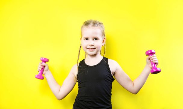 Theme sport and health. Beautiful caucasian child girl with pigtails posing on yellow background with smile. little athlete holding pink dumbbells. Banner for advertising, space for text copy space - Fotó, kép