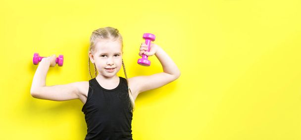 Theme sport and health. Beautiful caucasian child girl with pigtails posing on yellow background with smile. little athlete holding pink dumbbells. Banner for advertising, space for text copy space - 写真・画像