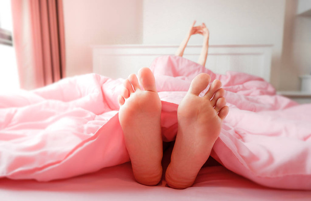 Foot in Bed at Home, Sleeping & Relax. Two Feet on the Bed in Pink Bedclothes. Beautiful Young Woman Barefoot in Bedroom Background. Female Feet is Lying fall Asleep on Pastel Bed Covered in Morning. - Φωτογραφία, εικόνα