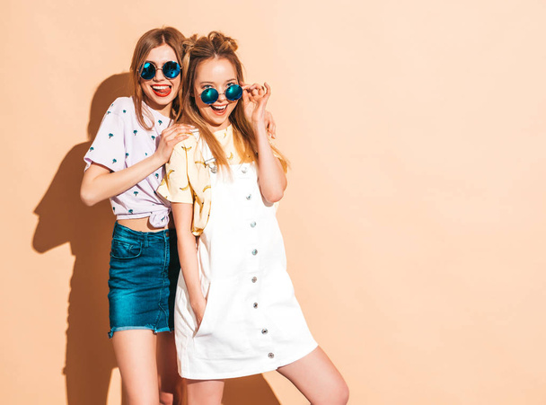 Two young beautiful smiling blond hipster girls in trendy summer colorful T-shirt clothes. Sexy carefree women posing near beige wall in round sunglasses. Positive models having fun - Фото, зображення