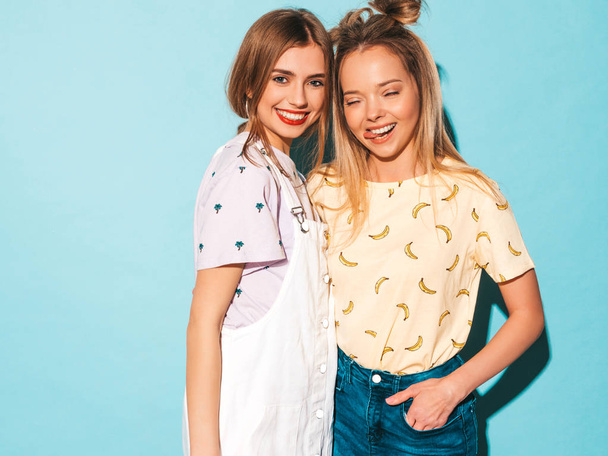 Twee jonge mooie glimlachend blond hipster meisjes in trendy zomer kleurrijke T-shirt kleding. Sexy zorgeloos vrouwen poseren in de buurt van blauwe muur. Positieve modellen plezier hebben en tonen van de tong - Foto, afbeelding