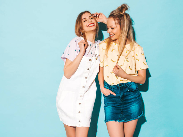 Two young beautiful smiling blond hipster girls in trendy summer colorful T-shirt clothes. Sexy carefree women posing near blue wall. Positive models having fun and hugging - Фото, зображення