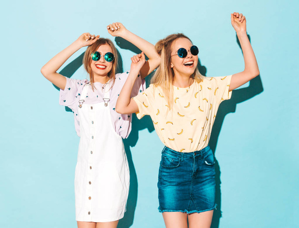 Two young beautiful smiling blond hipster girls in trendy summer colorful T-shirt clothes. Sexy carefree women dancing near blue wall in round sunglasses. Positive models having fun - Foto, Imagem