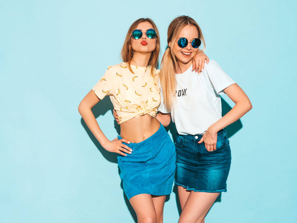 Two young beautiful smiling blond hipster girls in trendy summer colorful T-shirt clothes. Sexy carefree women posing near blue wall in round sunglasses. Positive models having fun - Foto, Bild