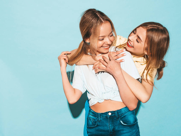 Две молодые красивые улыбающиеся блондинки-хипстерши в модной летней красочной одежде на футболке. Сексуальные беззаботные женщины позируют у голубой стены. Позитивные модели веселятся и обнимаются
 - Фото, изображение