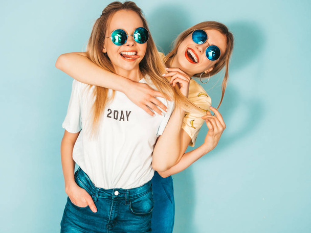 Две молодые красивые улыбающиеся блондинки-хипстерши в модных летних джинсах в юбках. Сексуальные беззаботные женщины позируют возле голубой стены в круглых солнцезащитных очках. Позитивные модели веселятся и обнимаются
 - Фото, изображение