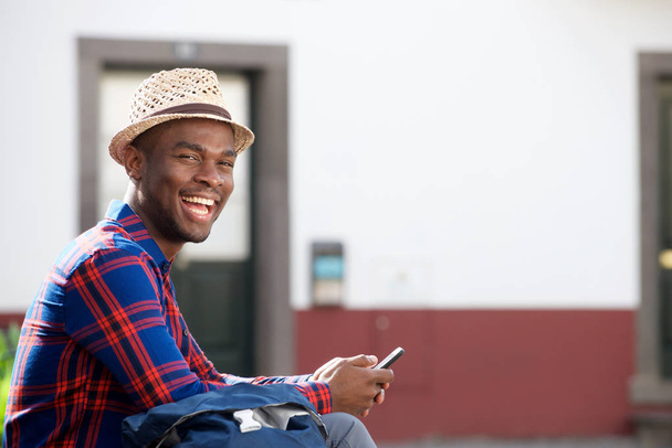 Portrait latéral de beau jeune homme afro-américain souriant avec téléphone portable à l'extérieur
 - Photo, image
