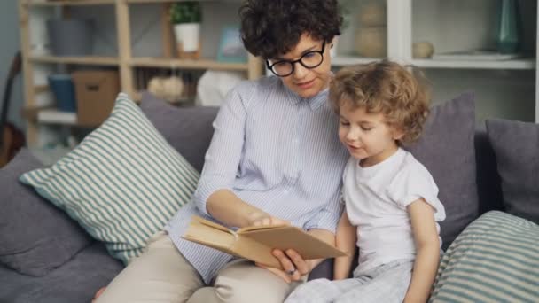 Loving mother teaching her son to read holding book speaking on couch at home - Video