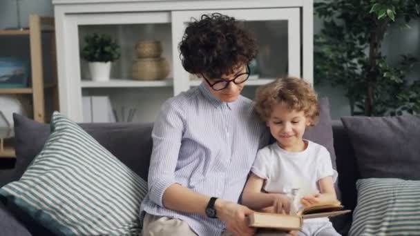 Happy mother and cute child reading book looking at pictures on sofa at home - Imágenes, Vídeo