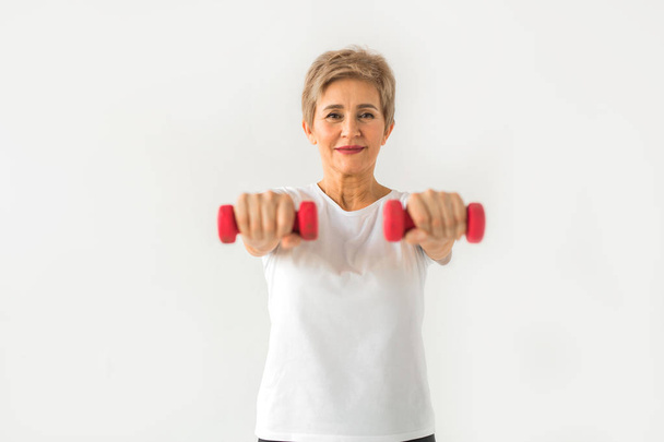stylish elderly woman in age stands playing sports with dumbbells on a white background - Foto, afbeelding