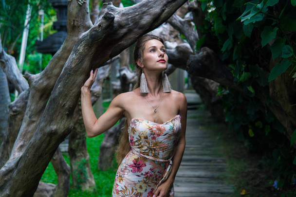 Beautiful young woman is leaning against a tree in the rain forest wearing sexy dress - Fotó, kép