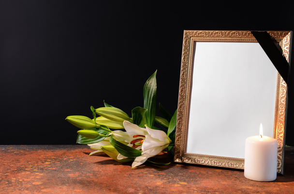 Blank funeral frame, candle and flowers on table against black background - Photo, image
