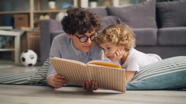 Young lady reading book to her son discussing story lying on floor at home - Filmmaterial, Video