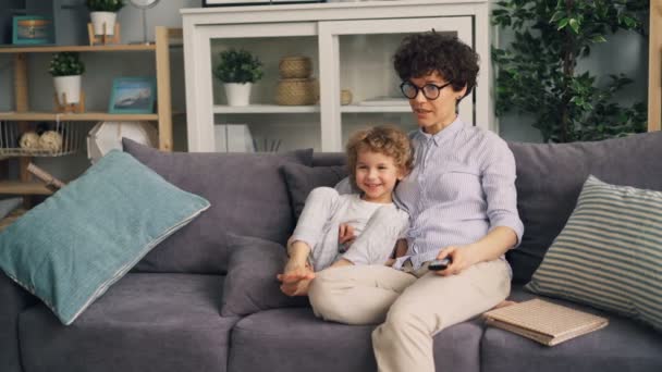 Mother and son watching TV at home talking and smiling sitting on sofa together - Materiał filmowy, wideo