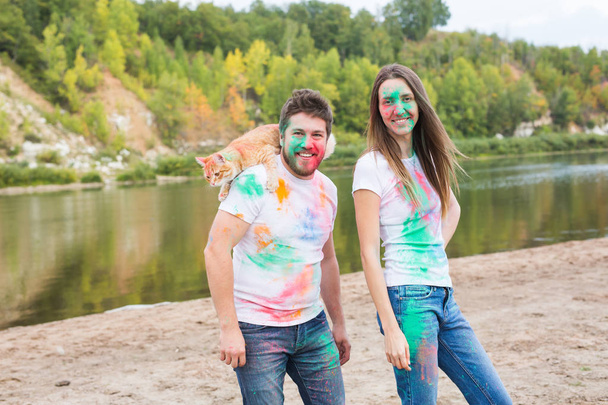 Festival holi, holidays, tourism and nature concept - Couple dressed in white shirts holding cat and covered with colorful dust - Valokuva, kuva