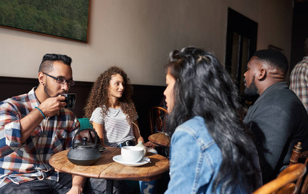 Diverse group of young friends sitting at tables in a trendy cafe drinking coffee and talking together - 写真・画像