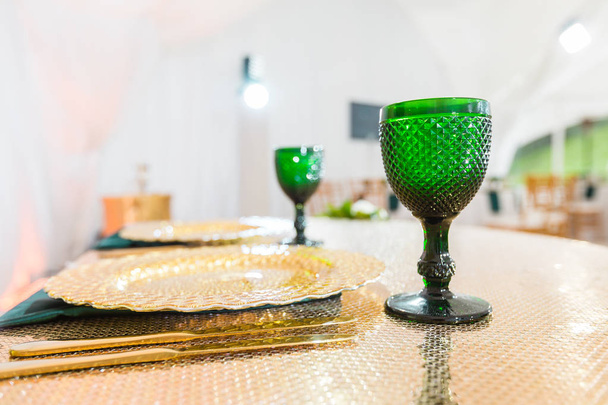 Gouden gerechten, groene wijnglazen en servetten. Horeca concept. Interieur van de tent voor bruiloft diner, klaar voor de gasten. Feestzaal tafel geserveerd. - Foto, afbeelding