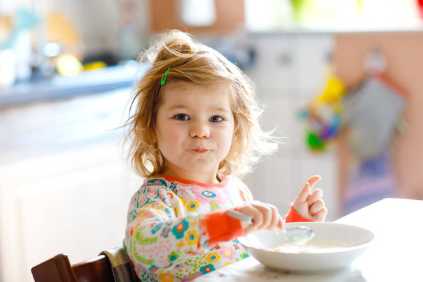 愛らしい幼児の女の子は、朝食のためにスプーンから健康的な粥を食べています。かわいいです幸せな赤ちゃん子供でカラフルなパジャマでキッチンに座ってスプーンを使用して学習. - 写真・画像