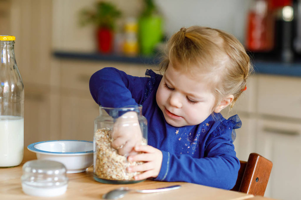 Adorable toddler girl eating healthy oatmeals with milk for breakfast. Cute happy baby child in colorful clothes sitting in kitchen and having fun with preparing oats, cereals. Indoors at home - Фото, изображение