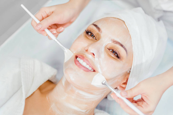 Cosmetology. The cosmetologist applies a cleansing face mask with two brushes. Smiling girl on the procedure for facial rejuvenation. Spa facial procedure - Photo, image