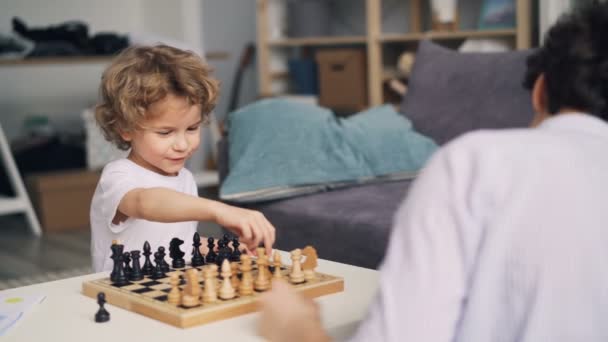 Joyful boy having fun with chess pieces while mom teaching him to play game - Imágenes, Vídeo
