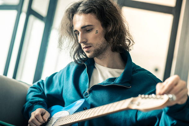 Melancholic long-haired young musician with small tattoo checking strings - Fotoğraf, Görsel