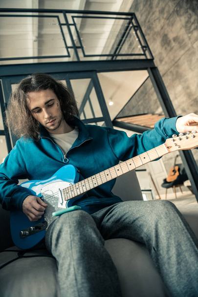 Dark-haired attentive man wearing blue sweatshirt and preparing guitar - Fotó, kép