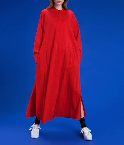 A young girl with blond long hair in a beautiful long red dress shirt, in white sneakers and black socks posing on a blue background, studio shot - Foto, afbeelding