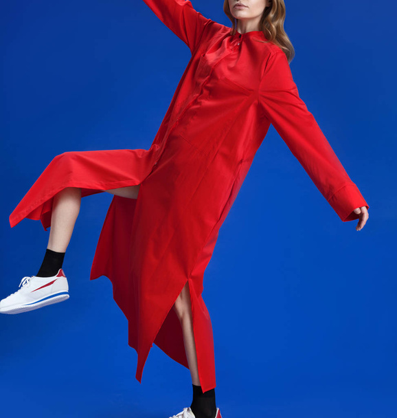 A young girl with blond long hair in a beautiful long red dress shirt, in white sneakers and black socks posing on a blue background, studio shot - Foto, imagen