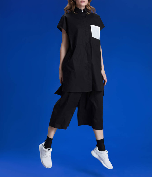 A young girl with blond long hair in a fashionable clothes, black culottes trousers, a black shirt, in white sneakers and black socks posing against a blue background, studio photography, jumping - Foto, imagen