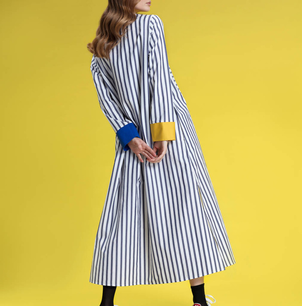 Young girl with long blonde hair in fashionable clothes, black and white with colored inserts long striped shirt dress posing on bright yellow background, studio shot - Foto, Imagem