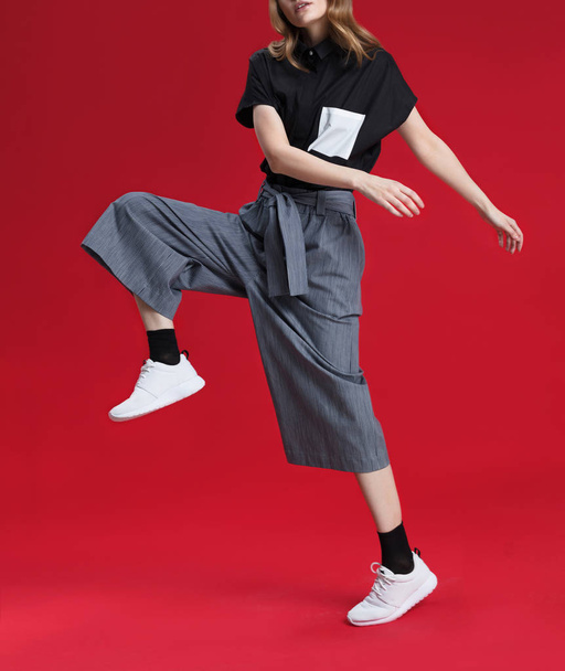 Beautiful blonde girl in gray trousers and black shirt, white sneakers, posing on red background, shooting in studio - Fotoğraf, Görsel