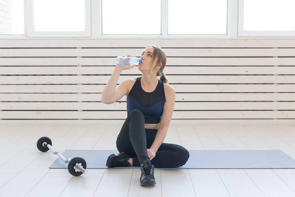 Фітнес, здорова і спортивна концепція - молода жінка сидить на схрещених ногах і п'є воду з пляшки після тренування
 - Фото, зображення