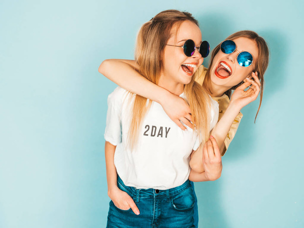 Two young beautiful smiling blond hipster girls in trendy summer jeans skirts clothes. Sexy carefree women posing near blue wall in round sunglasses. Positive models having fun and hugging - 写真・画像