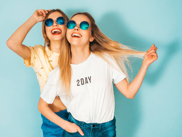 Two young beautiful smiling blond hipster girls in trendy summer jeans skirts clothes. Sexy carefree women posing near blue wall in round sunglasses. Positive models having fun and hugging - Foto, Imagem
