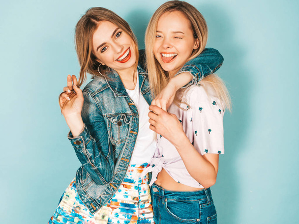 Two young beautiful smiling blond hipster girls in trendy summer colorful  clothes. Sexy carefree women posing near blue wall. Positive models having fun and showing tongue - 写真・画像